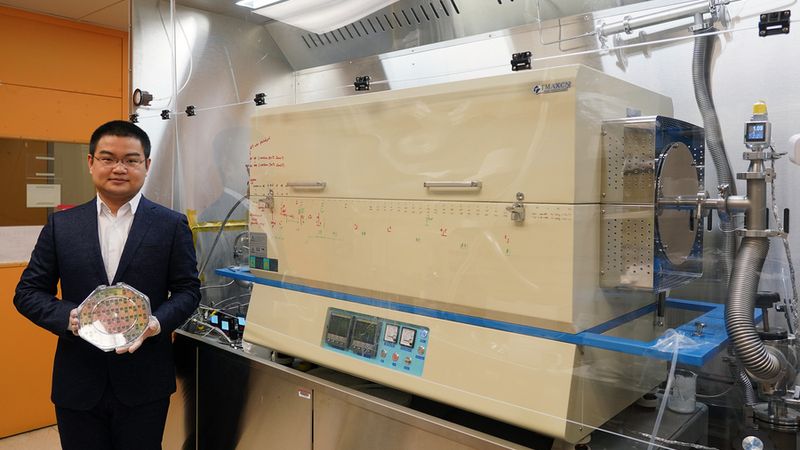 Graduate student Jiadi Zhu holding an 8-inch CMOS wafer with molybdenum disulfide thin film. On the right is the furnace the researchers developed, which enabled them to "grow" a layer of molybdenum disulfide onto the wafer using a low-temperature process that did not damage the wafer. Image: Courtesy of the researchers