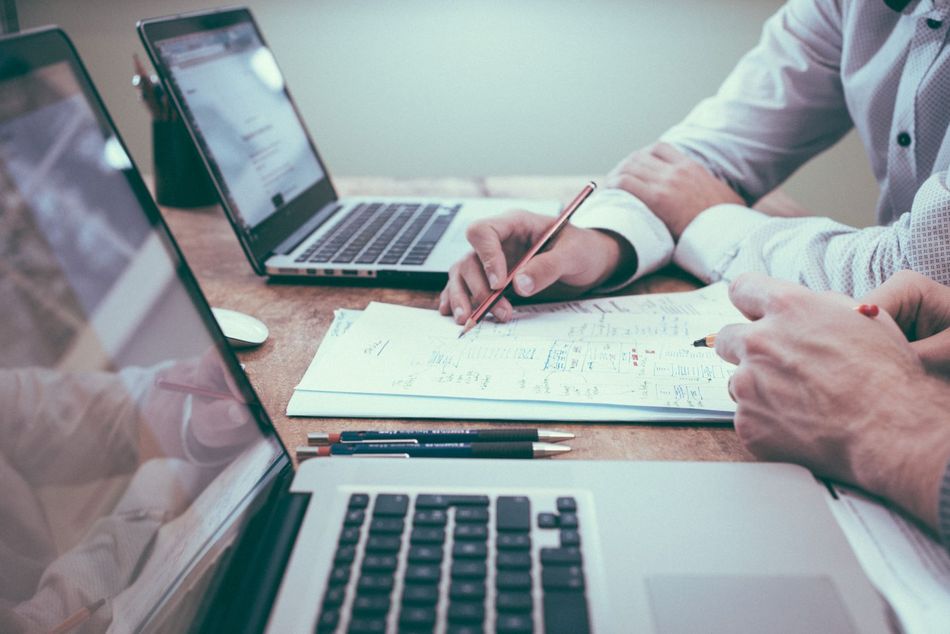 Two laptops sit next to each other, paper sits between them. Two people's hand are seen writing together on the paper. 