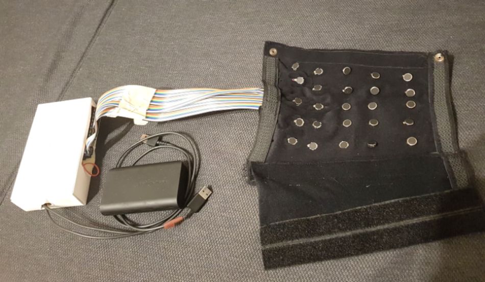 A picture of a custom-built haptic feedback sleeve, comprised of a grid of 25 vibration motors sewn onto fabric. Wires are seen leading to a control box and a battery pack.