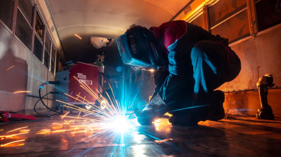 A photograph of a welder using a metal inert gas (MIG) welder to attach steel sheets to the floor of a bus from the inside.