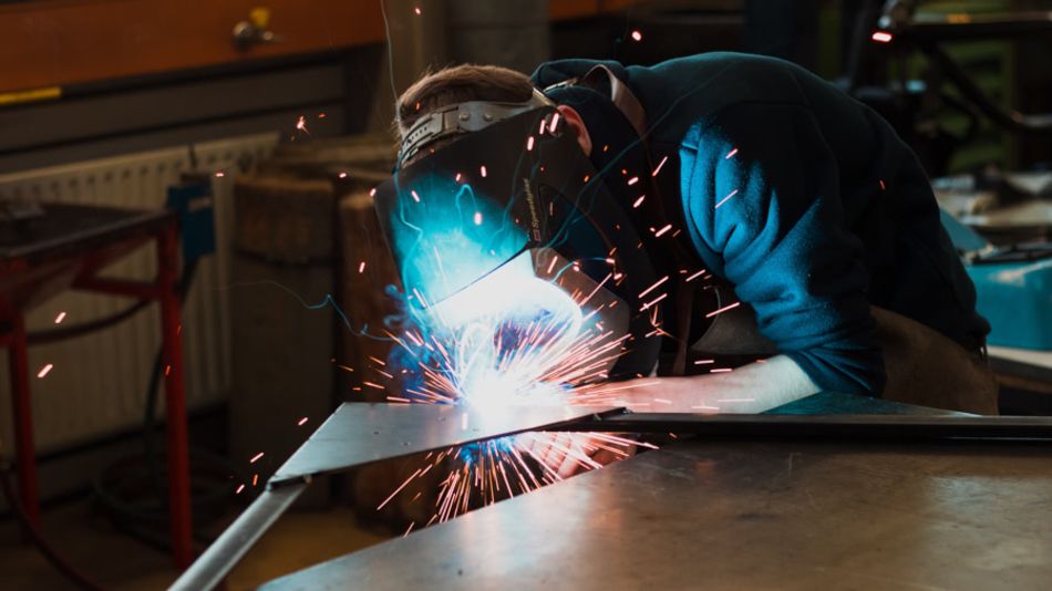 A person in a welding mask using a welding gun to attach sheet metal to metal tubes; sparks can be seen flying.