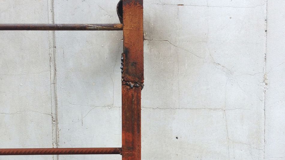 A photograph of a metal ladder against a white wall. The ladder has been repaired by welding, with an ugly weld visible on the right-hand string.