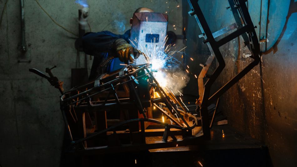 A photograph of a welder, seen from the front, working on a large metal object; a hand-held welding mask is used to protect the welder's eyes from the arc.