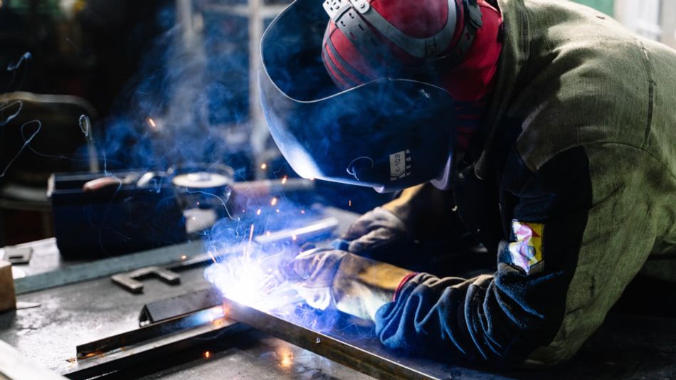 An image of a welder attaching metal bar; a welding mask is worn on the head and the welding gun's arc is visibly sparking.