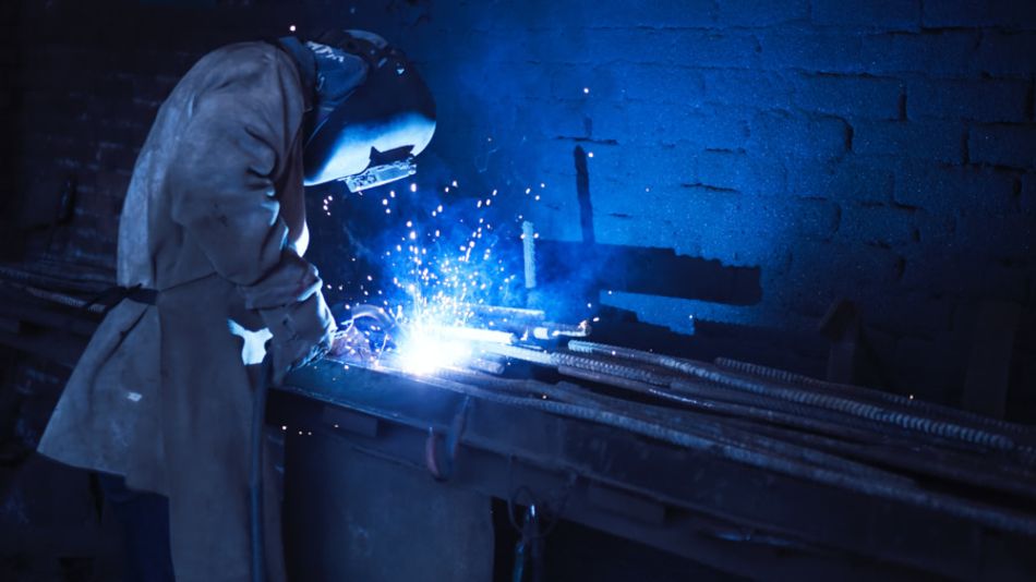 A photograph of a welder working on metalwork in a dimly-lit room. Protective coveralls are worn along with a welding mask, and sparks can be seen flying from the welding gun's tip.