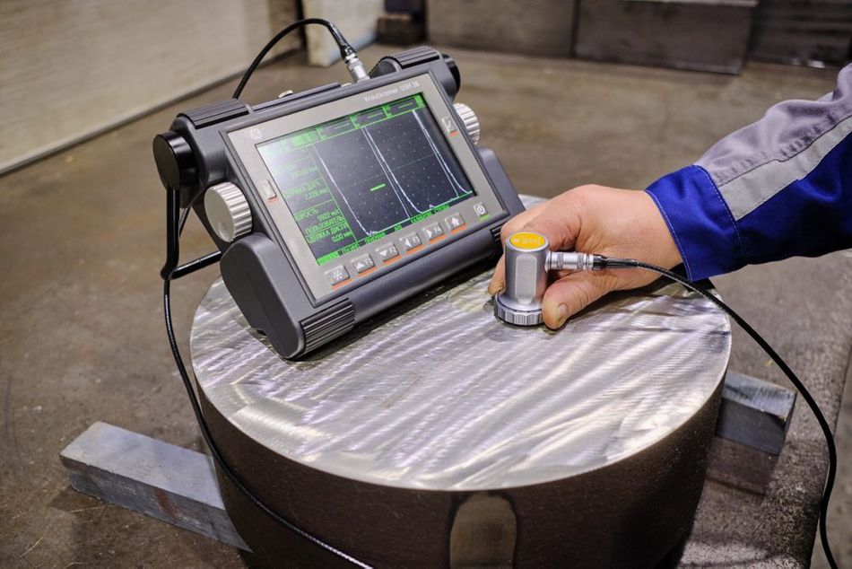 A person's hand holding an ultrasonic thickness gauge probe on a metal surface, next to a portable ultrasonic flaw detector displaying a graph.