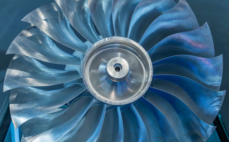 A shiny metal turbine with multiple blades is centered on a circular hub made of titanium, set against a blue background in a close-up view.