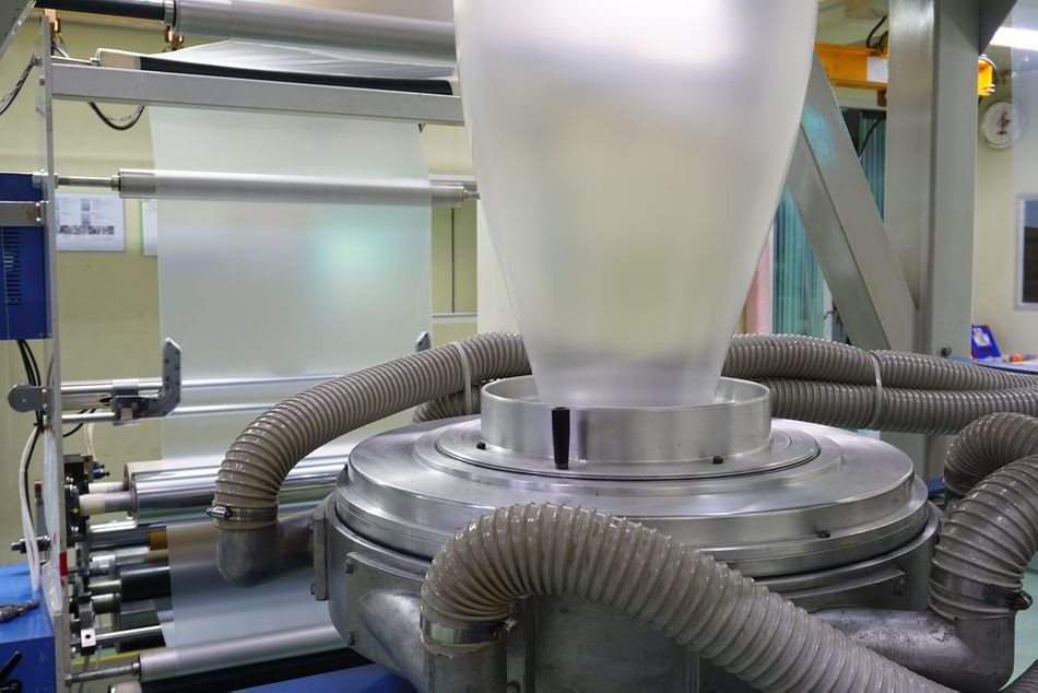 A detailed view of a plastic extrusion machine used in a manufacturing facility. The machine is forming a large, thin film bubble from molten plastic, with metal rollers and hoses visible. The background shows additional machinery and equipment involved in the production process.