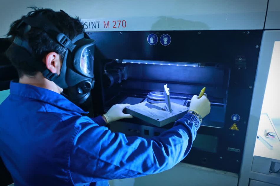 A person wearing a protective mask and gloves is operating a metal 3D printing machine. The person is examining a 3D-printed part inside the machine. The scene is well-lit, highlighting the detailed and technical environment of the 3D printing process.