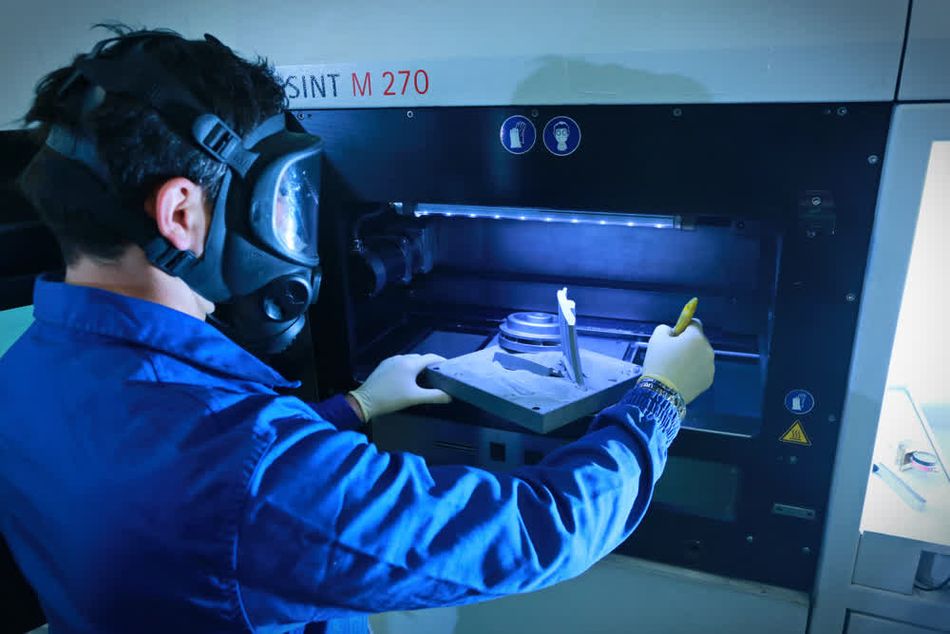 A person wearing a protective mask and gloves is operating a metal 3D printing machine. The person is examining a 3D-printed part inside the machine. The scene is well-lit, highlighting the detailed and technical environment of the 3D printing process.