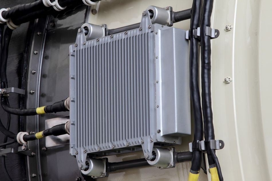 A close-up image of a large, gray metal box with cooling fins mounted on a beige wall, with black cables connected and secured by cable ties.