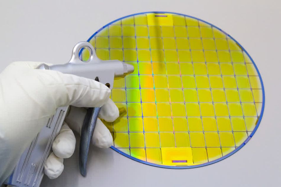 A close-up image illustrating the wafer cleaning process in the semiconductor industry, emphasizing the integration of plasma cleaning and nitrogen-based techniques. A gloved hand holds a specialized nitrogen gun, directing a controlled stream of nitrogen gas over a highly reflective silicon wafer. The wafer features a grid of microcircuits and exhibits an iridescent yellow and blue sheen, characteristic of its optical coatings and precision-engineered surface. This image highlights the critical role of nitrogen cleaning in removing particulate contaminants and residues, complemented by plasma cleaning to ensure a pristine surface. Both techniques are essential in maintaining the integrity of the wafer for advanced semiconductor fabrication processes. The use of gloves underscores the importance of cleanroom standards in preventing contamination.