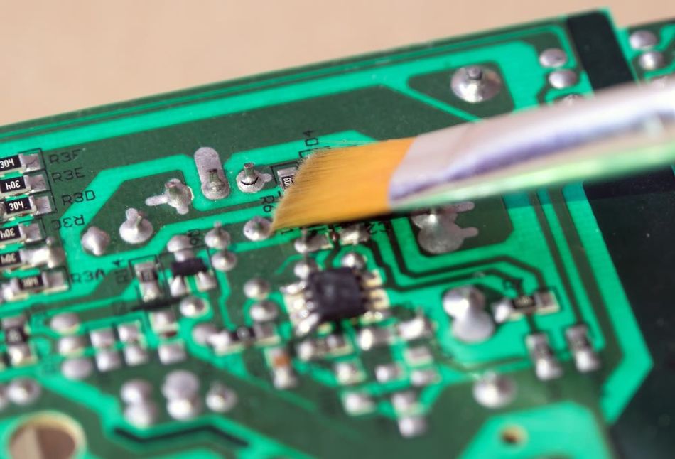 Close-up of a printed circuit board (PCB) being cleaned with a soft brush, showing multiple solder joints and electronic components. The image highlights attention to solder quality and maintenance practices, including addressing potential cold solder joints or flux residue.