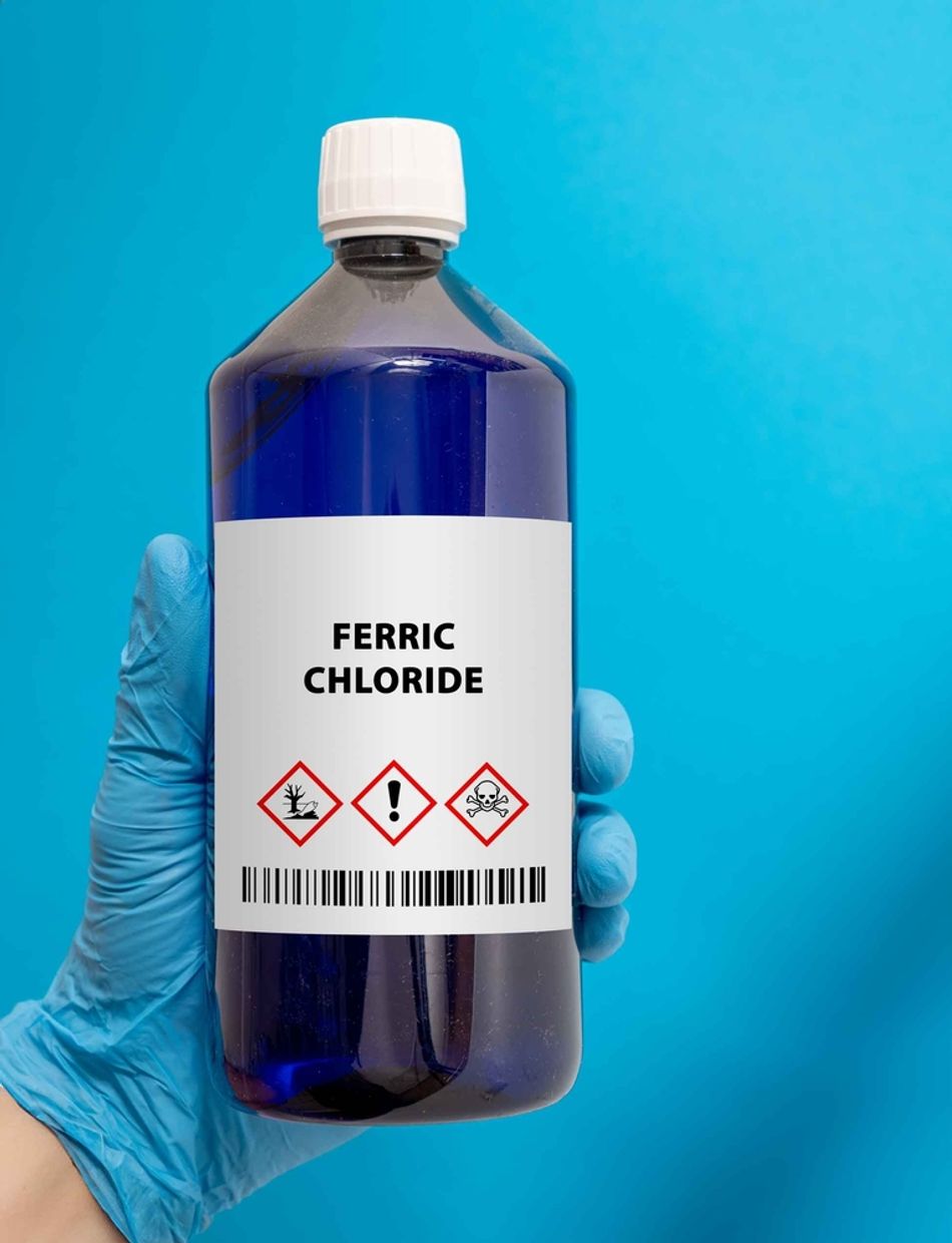 A gloved hand holds a labeled bottle of ferric chloride, a chemical etchant commonly used in printed circuit board (PCB) manufacturing to remove unwanted copper during the etching process. The bottle features hazard symbols, emphasizing the need for careful handling in electronics fabrication.